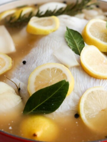 A whole chicken submerged in brine with lemon slices, bay leaves, onion, and herbs in a pot.
