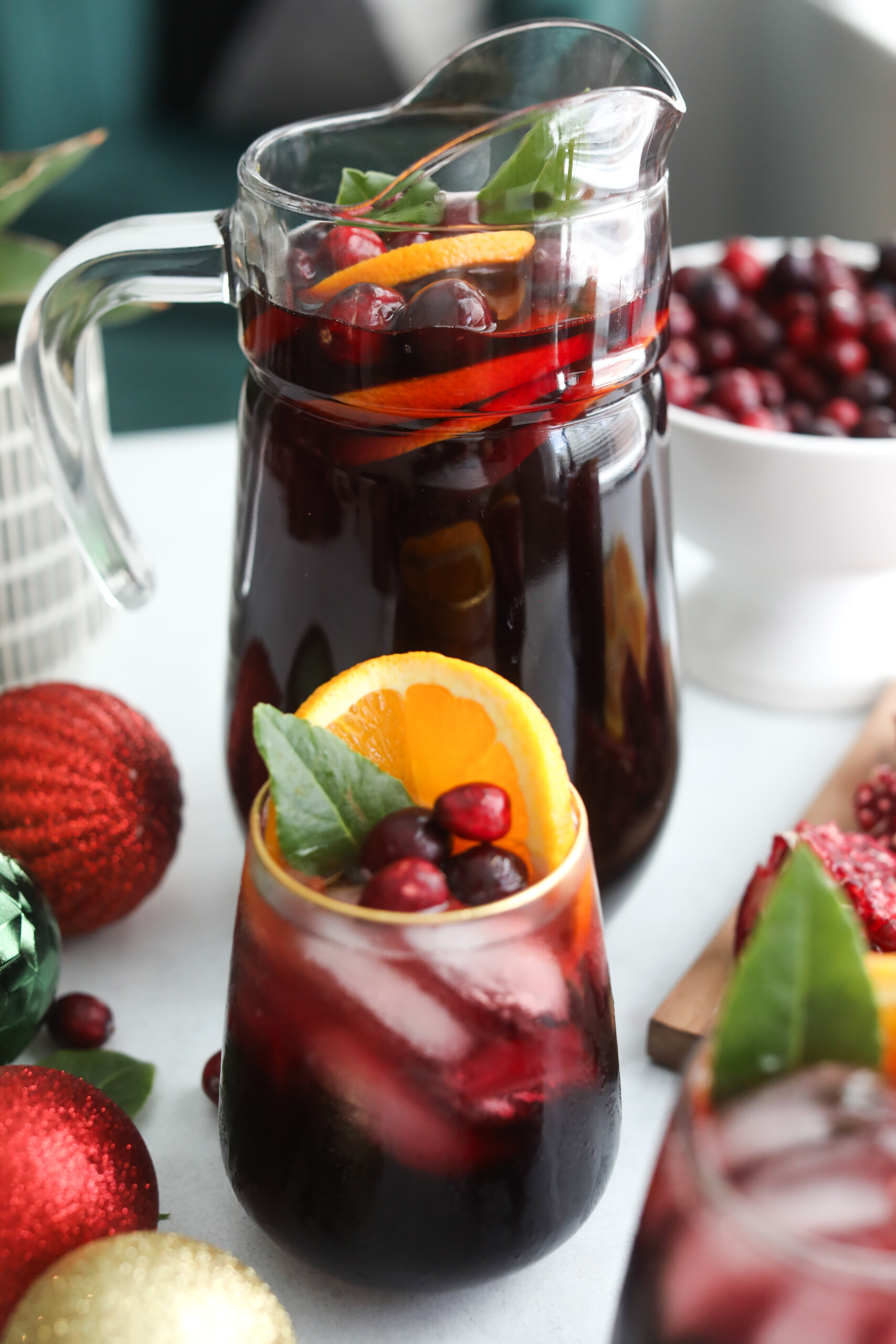 Christmas punch recipe Glass pitcher and wine glass filled with deep red Christmas Punch, garnished with orange slices, cranberries, and green leaves, surrounded by festive ornaments.