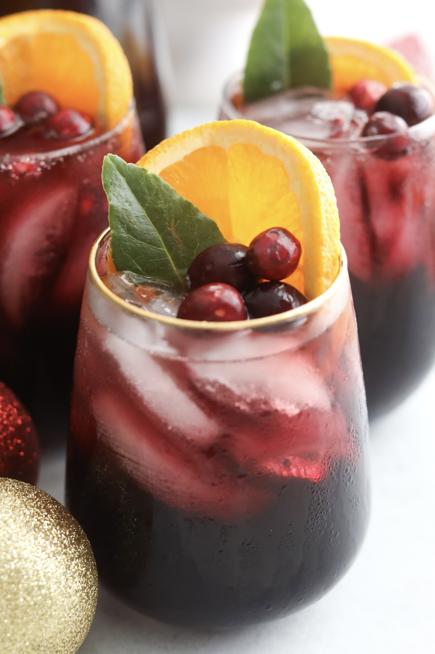 Close-up of a chilled glass of Christmas Punch showing condensation on the outside and garnishes of orange, cranberries, and green leaves.