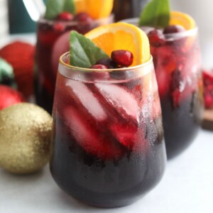 Close-up of a glass of Christmas Punch with condensation, filled with ice and topped with orange slices, cranberries, and a green leaf.