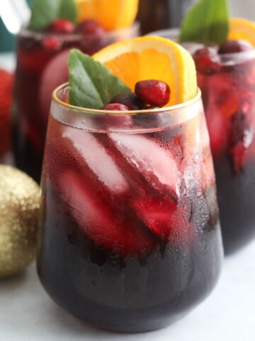 Close-up of a glass of Christmas Punch with condensation, filled with ice and topped with orange slices, cranberries, and a green leaf.