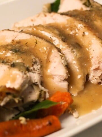 Sliced slow cooker turkey breast arranged on a platter with gravy, carrots, and herbs.