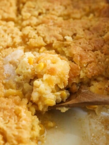 a close-up scoop of soft, golden cornbread pudding being lifted from the baking dish with a wooden spoon.