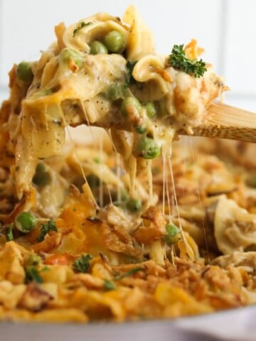 A close-up of a wooden spoon lifting a cheesy scoop of turkey casserole filled with peas, noodles, and crispy fried onions.