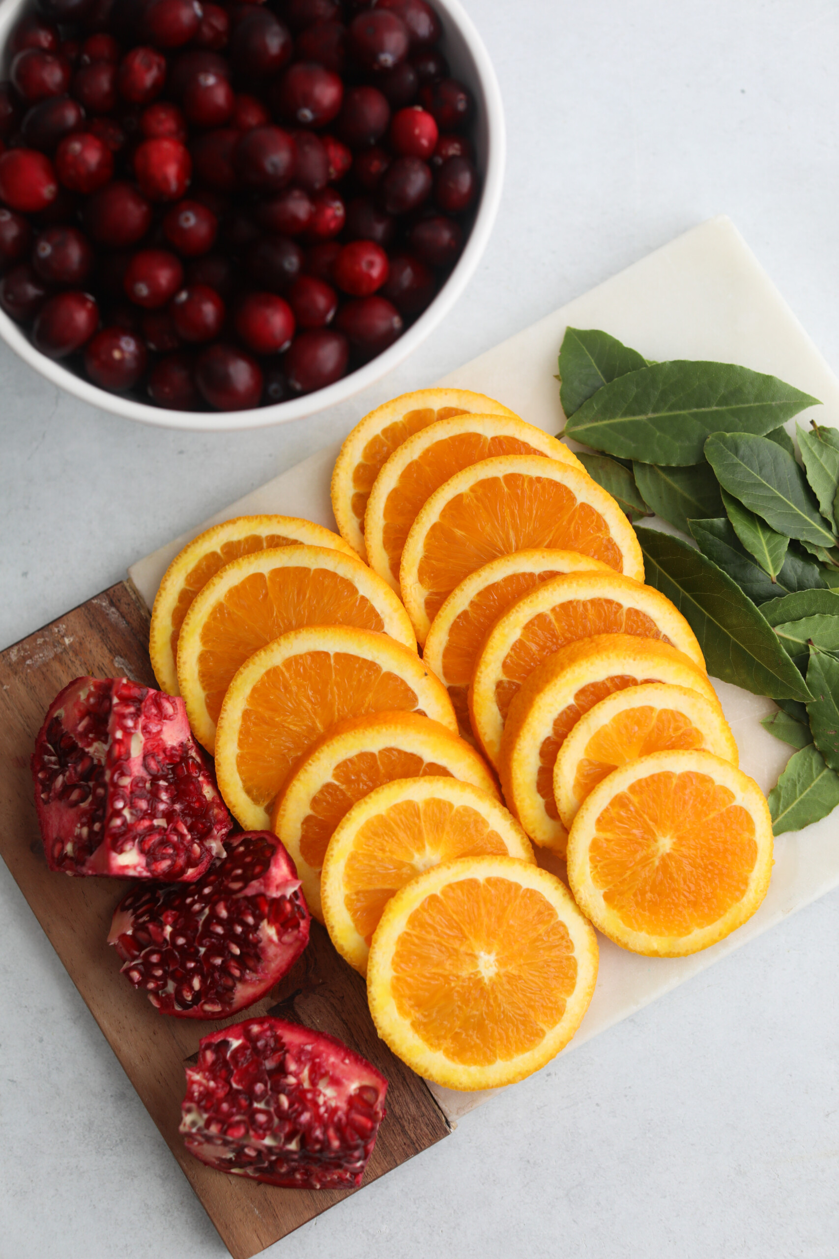 Sangria for the holidays Overhead view of a cutting board topped with orange slices, pomegranate pieces, and bay leaves beside a white bowl of cranberries.