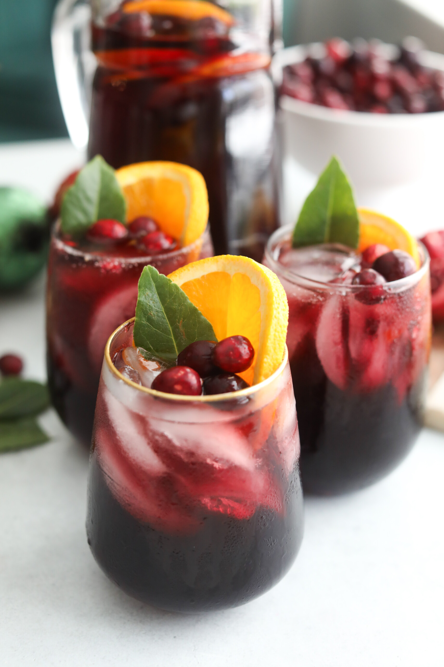 Holiday sangria Three glasses of chilled Christmas Punch with orange slices, cranberries, and greenery in front of a pitcher of punch and a bowl of fresh cranberries.